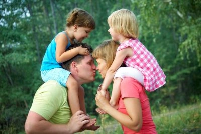 glückliche familie - Ehepaar mit jeweils einem Kind auf den Schultern am Waldrand, gibt sich einen Kuss